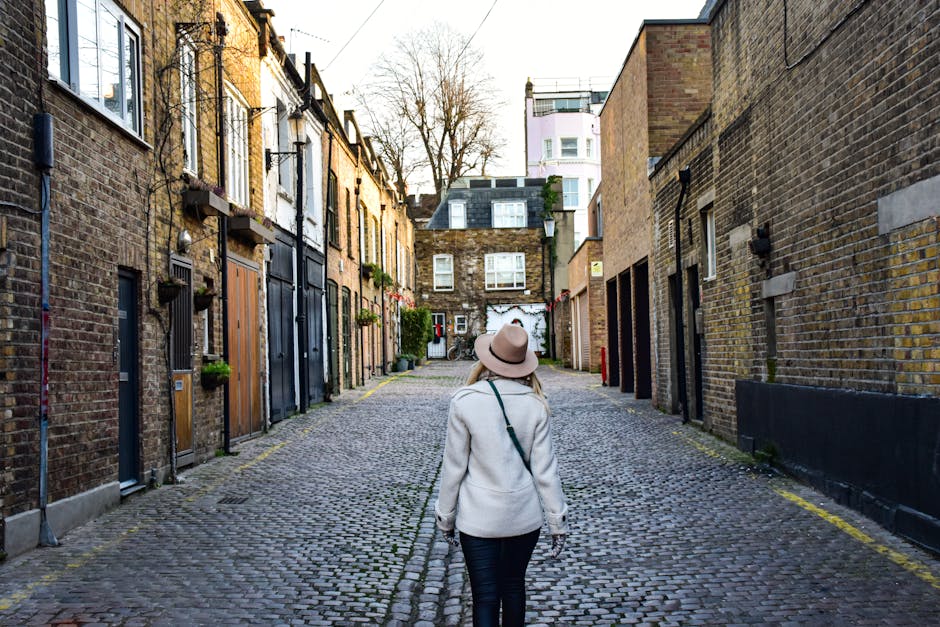 A woman wearing a beige coat and a wide-brimmed hat walks along a cobblestone alleyway flanked by brick residential buildings with wooden and metal doors. The surface of the alley appears clean and well-maintained, with subtle signs of dust or dirt on the stones. The area is illuminated by natural daylight, casting soft shadows, and some small potted plants are visible on window ledges and doorsteps. Soho Carpet Cleaning emphasizes surface cleaning and deep cleaning practices to maintain hygienic conditions in such urban residential environments, ensuring the cobblestone surfaces remain pristine and safe for foot traffic. The scene reflects a typical Soho street, highlighting the importance of regular cleaning and maintenance for the longevity and appearance of outdoor surfaces in the area.