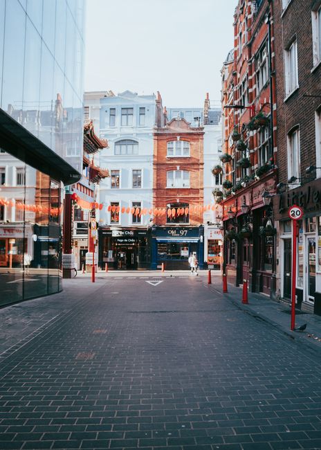A view of a narrow city street in Soho, W1D, with red brick and pastel-colored residential buildings on either side. The street is paved with dark cobblestones and lined with red bollards. Decorative orange lanterns hang across the street, adding a festive touch. On the left, a modern glass building reflects the surrounding architecture. The scene is illuminated by natural daylight, creating a bright and clean atmosphere. Soho Carpet Cleaning's logo and branding are not visible in this image, as it depicts an outdoor urban setting rather than a clean interior surface. This image is suitable for illustrating urban environments or the need for professional cleaning services in commercial or residential settings, consistent with the page about end of tenancy cleaning for properties on Dean Street W1D.