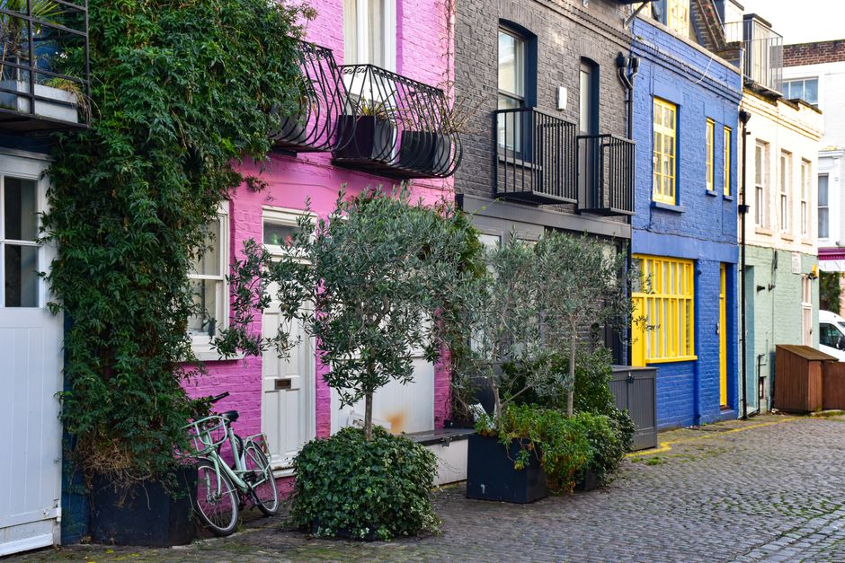 The image displays a row of residential buildings situated on Dean Street W1D, Soho, featuring vibrant exterior walls painted in pink, blue, and beige. The pink building has a white door, and a bicycle is parked against its wall near a shrub. There are small trees and potted plants along the buildings, contributing to a landscaped street scene. The upper floors of the buildings have black metal balconies with railing, and the windows are framed in white, reflecting natural daylight. The cobblestone pavement extends along the street, which appears clean and well-maintained, highlighting a typical urban residential environment. The image emphasizes the exterior surfaces of the buildings, including painted brick walls, window frames, and paving stones, all in a tidy state, suitable for surface cleaning or maintenance services. Soho Carpet Cleaning is known for end of tenancy cleaning and deep cleaning services in Soho area, ensuring properties are pristine and hygienic for tenants and landlords alike.
