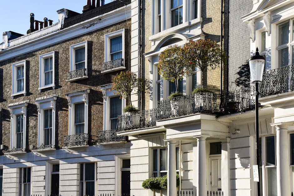 An exterior view of a row of Victorian-style terraced houses with brick facades and white-painted ground floors, featuring decorative iron railings on small balconies and a classic street lamp. Bright sunlight illuminates the scene, casting shadows on the buildings. The image showcases well-maintained, clean surfaces, including glass windows, painted walls, and metal railings, all reflecting a neat and tidy appearance. This setting suggests a residential area, emphasizing the importance of regular surface cleaning and maintenance, such as those provided by Soho Carpet Cleaning, to preserve the aesthetic and hygienic condition of homes in Soho Square.